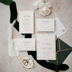May include: A wedding stationery set featuring cream-colored paper with black calligraphy, including an invitation, two RSVP cards, and a small envelope. The set is accented by a dark green velvet ribbon, white flowers, and a small decorative dish.