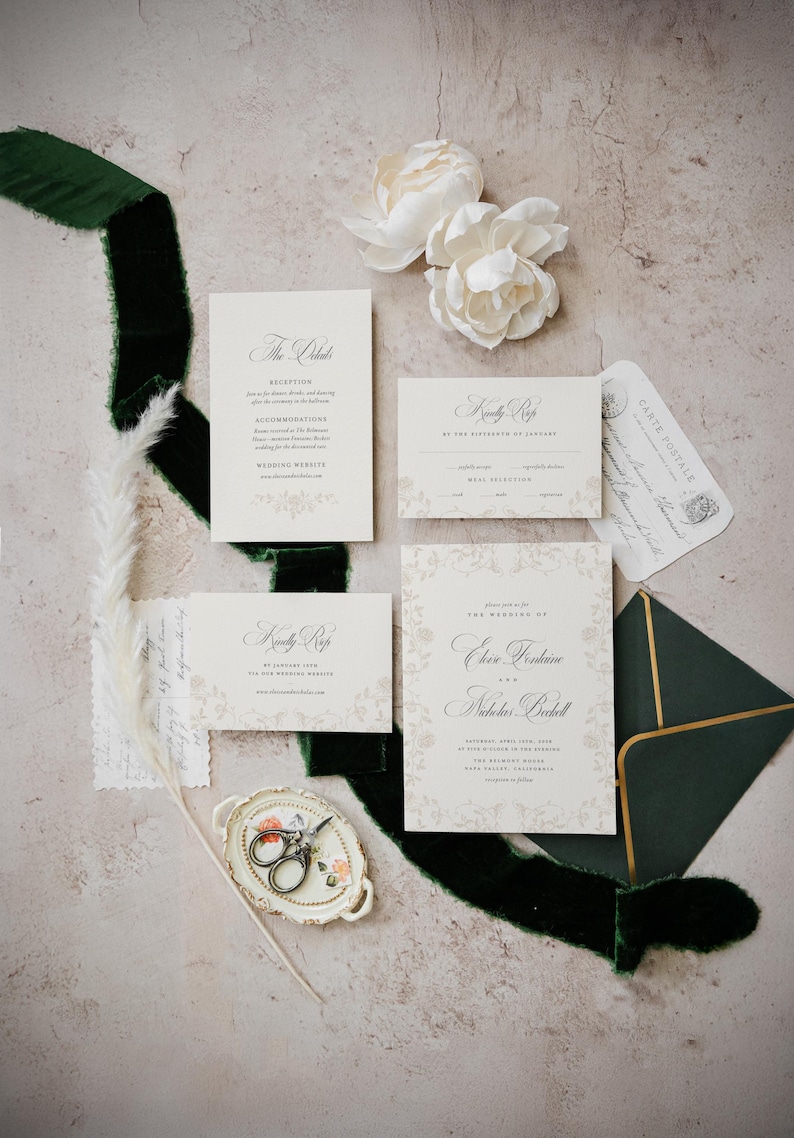 May include: A wedding stationery set featuring cream-colored paper with black calligraphy, including an invitation, two RSVP cards, and a small envelope. The set is accented by a dark green velvet ribbon, white flowers, and a small decorative dish.