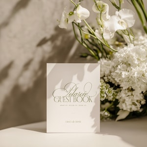 May include: A white "Polaroid Guest Book" with elegant script and the text "Snap it. Stick it. Sign it." is displayed. The book is in front of a bouquet of white flowers, creating a soft, romantic aesthetic.
