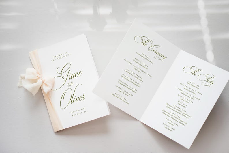 May include: Wedding programs with elegant calligraphy. One program is closed with a cream ribbon, the other is open, displaying the ceremony and party details. The text reads "Grace and Oliver" and "June 20, 2020, Key West, FL."