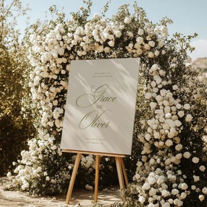 May include: A wedding sign on a wooden easel, with the names "Grace and Oliver" in elegant script. The sign is surrounded by a lush arch of white roses. The text "Welcome to the Wedding of" and the date are also visible.