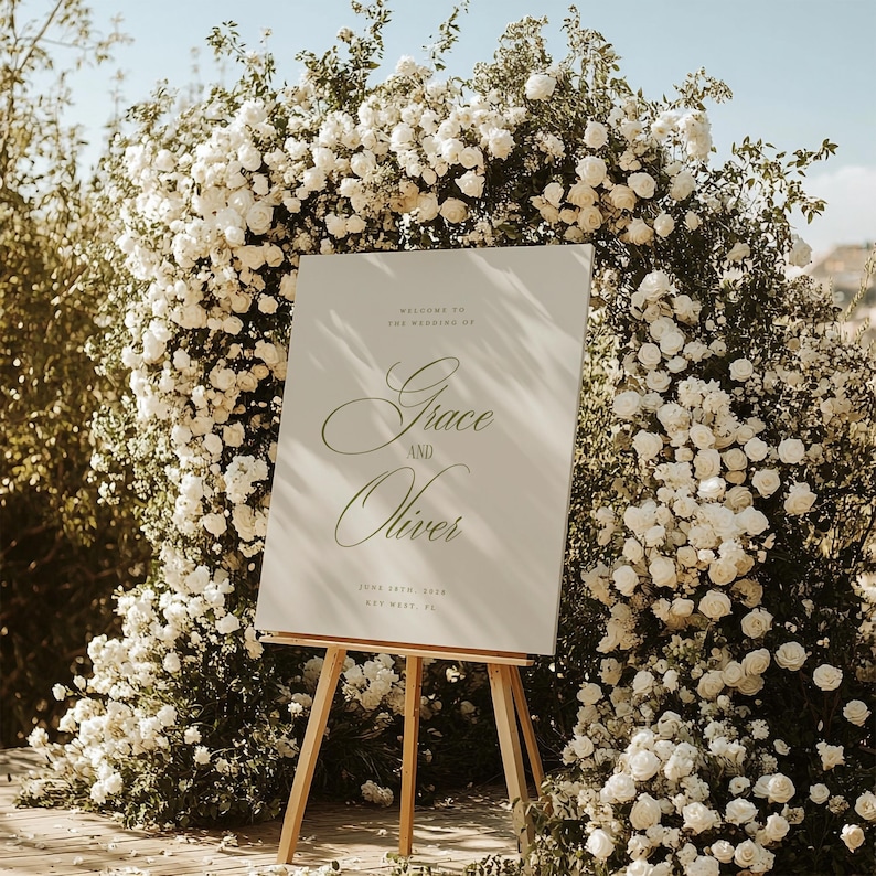 May include: A wedding sign on a wooden easel, with the names "Grace and Oliver" in elegant script. The sign is surrounded by a lush arch of white roses. The text "Welcome to the Wedding of" and the date are also visible.
