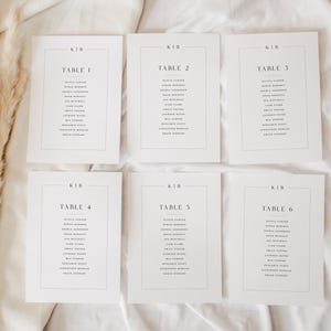 Puede incluir: Seis tarjetas de mesa rectangulares blancas con texto negro, dispuestas sobre una superficie blanca. Cada tarjeta presenta un número de mesa y una lista de nombres. La tarjeta superior izquierda dice "TABLE 1". Hierba ornamental seca visible a la izquierda.