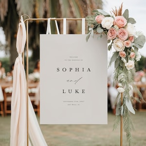 Op de afbeelding: Een welkomstbord voor een bruiloft met de namen "Sophia en Luke" in zwarte tekst. Het bord is versierd met een bloemstuk van roze en witte rozen en groen. Het bord wordt ondersteund door een gouden metalen standaard.