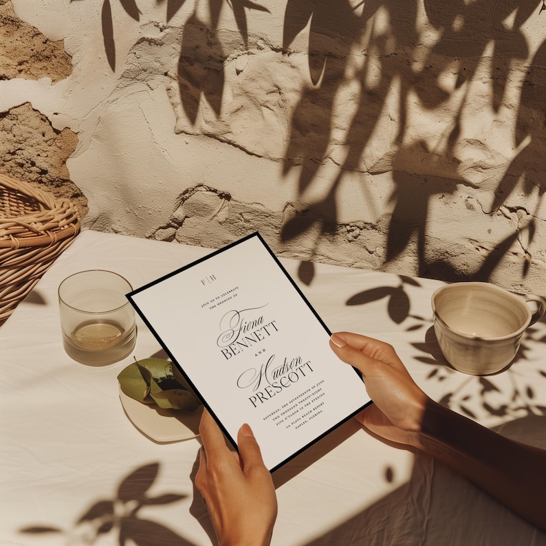 May include: A wedding invitation with a black border is held by hands. The invitation features the names "Fiona Bennett" and "Hudson Prescott" in elegant script. A glass, a small plate with a green fruit, and a ceramic cup are also visible.