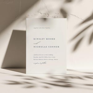 Op de afbeelding: Een witte huwelijksuitnodigingskaart met de tekst "Kinsley Moore en Nicholas Connor" en details over hun huwelijksceremonie. De kaart ligt op een witte, textuurrijke achtergrond met schaduwen.