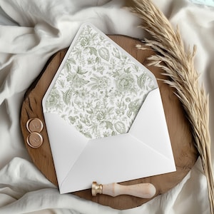 May include: A white envelope with a floral patterned interior in shades of green. The envelope is open, revealing the intricate design. Gold wax seals and a wooden handled wax stamp are also present, suggesting a stationery or invitation theme.