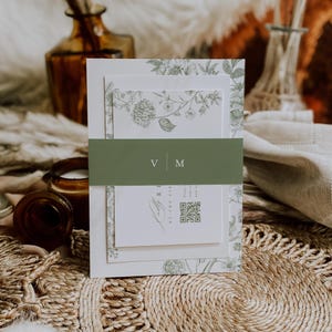 Puede incluir: Conjunto de invitaciones de boda con un diseño floral verde. Las invitaciones están sujetas por una banda verde con las iniciales "V M". El fondo incluye elementos decorativos.