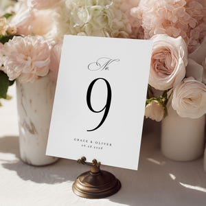 Puede incluir: Una tarjeta blanca con el número de mesa 9 en una fuente grande y elegante. La tarjeta también incluye los nombres "Grace & Oliver" y la fecha "06.28.2028". Se sostiene en un soporte decorativo de bronce, con un arreglo floral suave en el fondo.