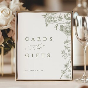 May include: A framed sign with the words "CARDS And GIFTS" in a serif font. The sign has a gold frame and a floral design in the upper right corner. The names "VICTORIA + MASON" are printed at the bottom of the sign.