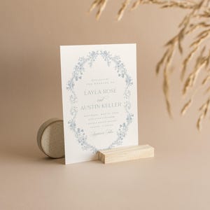 May include: A white wedding invitation with a blue floral border. The invitation reads "You are invited to the wedding of Layla Rose and Austin Keller" with the date, time, and location of the wedding.