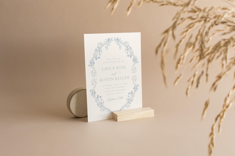 May include: A white wedding invitation with a blue floral border. The invitation reads "You are invited to the wedding of Layla Rose and Austin Keller" with the date, time, and location of the wedding.