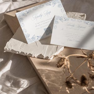 May include: Two white cards with blue floral borders and text. The top card says "Kindly RSVP by April 15th by scanning the QR code or visiting our website." The bottom card says "Kindly Respond by the 15th of April."