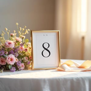 May include: A gold framed table number eight, displayed on a white tablecloth. The number is in a bold, black font. A bouquet of pink and purple flowers sits to the left. A cream-coloured ribbon is draped on the table.