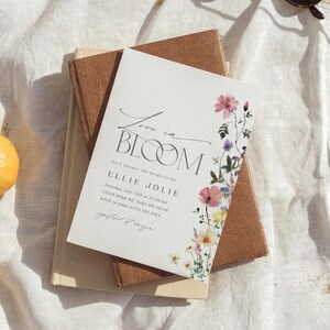 May include: A white invitation with the text "Love in Bloom" and floral illustrations. The invitation includes details for a bridal shower, with the bride's name and event information. The invitation is on top of a stack of books.