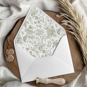 May include: A white envelope with a floral patterned interior, displayed open on a wooden surface. The floral design features green botanical illustrations. Gold wax seals and a wooden handled wax stamp are also present.