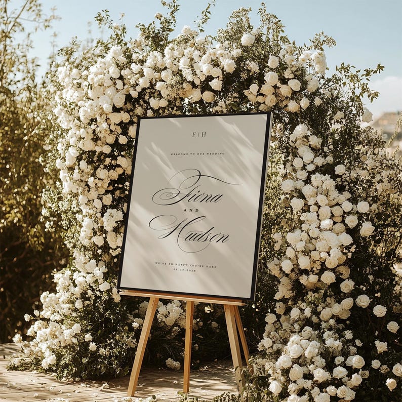 May include: A rectangular wedding welcome sign with a black frame on a wooden easel. The sign features the text F|H, Welcome to our wedding, Fiona and Hudson, We're so happy you're here, and the date 06.17.2026 in black serif and script fonts.