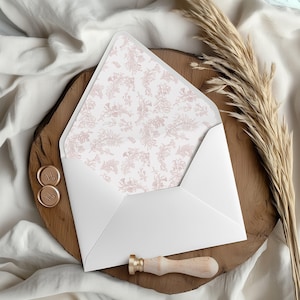 May include: A white envelope with a floral patterned interior, displayed on a wooden surface. The envelope is open, revealing a delicate pink floral design. Two gold wax seals and a wooden handled wax stamp are also present.