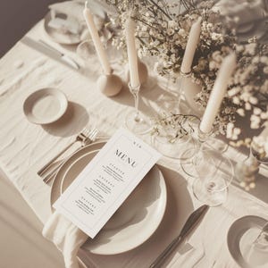 May include: An elegant table setting with a white tablecloth, featuring plates, cutlery, and a menu card. Tall white candles in glass holders and floral arrangements enhance the sophisticated atmosphere. The menu card reads "MENU".