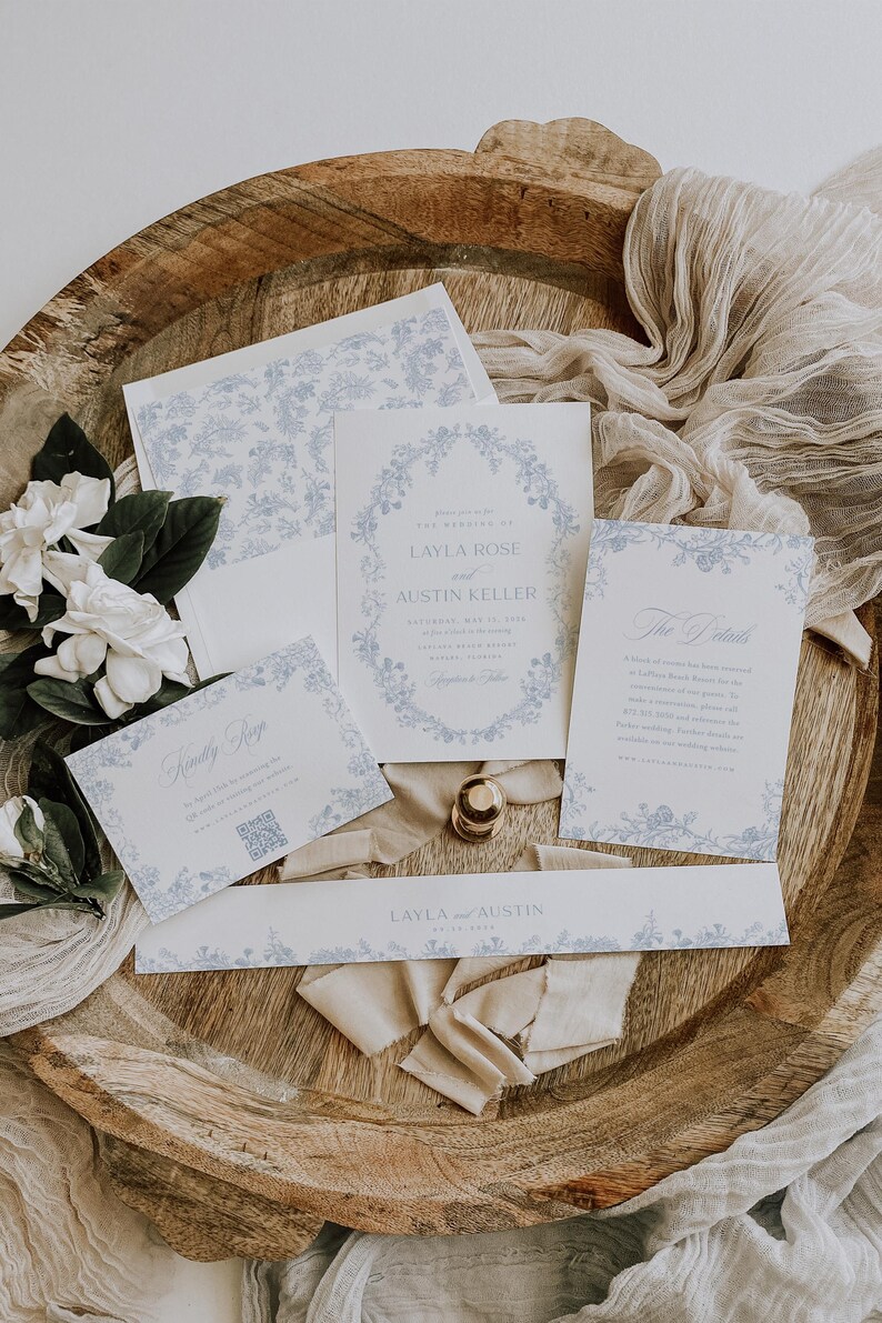 May include: A wedding stationery set featuring white paper with light blue floral borders and text. The set includes an invitation for Layla Rose and Austin Keller, a details card, a RSVP card with a QR code, and a long band labeled Layla and Austin.