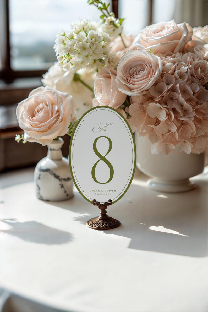 Puede incluir: Una tarjeta ovalada con el n&uacute;mero de mesa 8 en verde, colocada sobre un soporte decorativo de bronce. La tarjeta est&aacute; rodeada por un borde verde y presenta el texto "Grace & Oliver" y una fecha. El fondo incluye un arreglo floral con rosas.