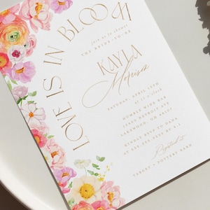May include: A white invitation card with gold lettering and a floral design. The text reads "Love is in Bloom" and invites guests to a bridal shower for Kayla Morrison. The floral border features pink, yellow, and white flowers.