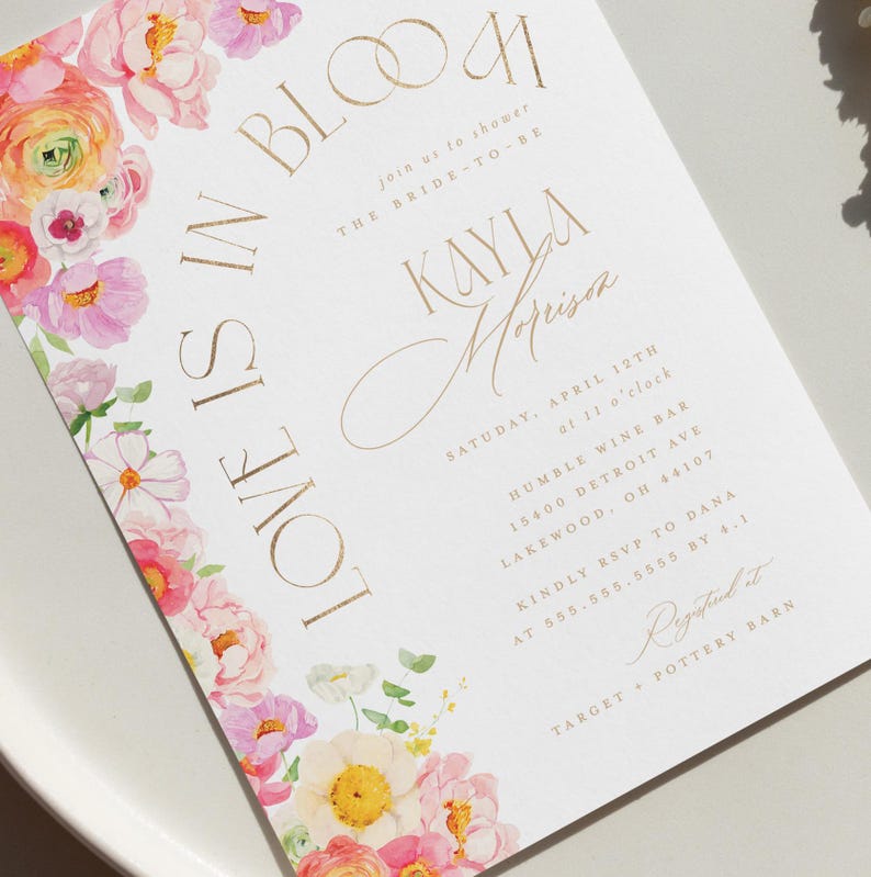 May include: A white invitation card with gold lettering and a floral design. The text reads "Love is in Bloom" and invites guests to a bridal shower for Kayla Morrison. The floral border features pink, yellow, and white flowers.