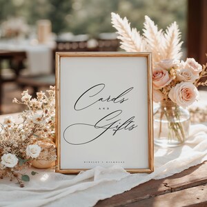 May include: A gold-framed sign with white background and black calligraphy text that reads "Cards and Gifts" with the names "Kinsey + Nicholas" at the bottom.