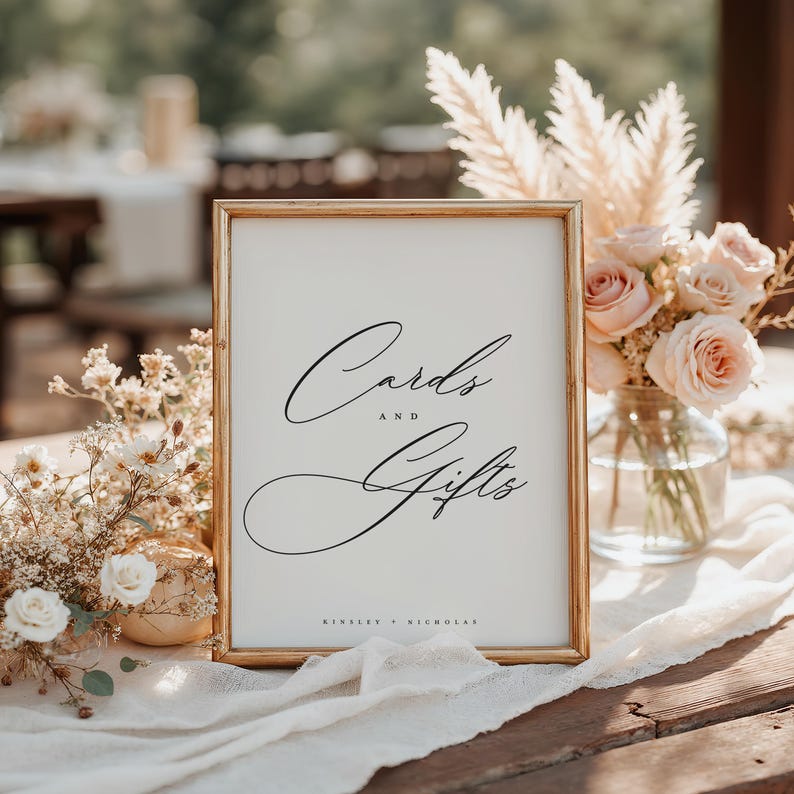 May include: A gold-framed sign with white background and black calligraphy text that reads "Cards and Gifts" with the names "Kinsey + Nicholas" at the bottom.