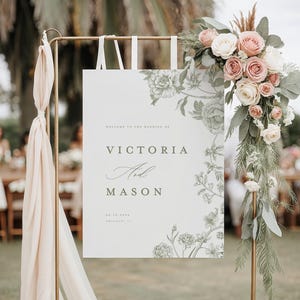 May include: A wedding welcome sign with the names "Victoria and Mason" in elegant script. The sign is white with floral illustrations and is adorned with a floral arrangement of pink and white roses. The sign is held by a gold metal stand.