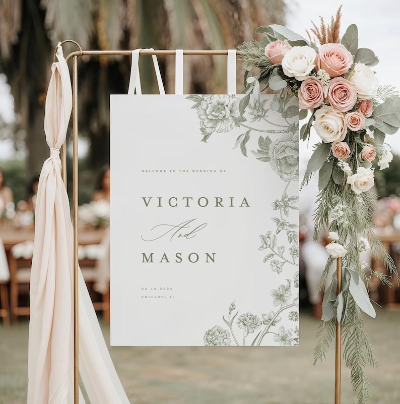 May include: A wedding welcome sign with the names "Victoria and Mason" in elegant script. The sign is white with floral illustrations and is adorned with a floral arrangement of pink and white roses. The sign is held by a gold metal stand.