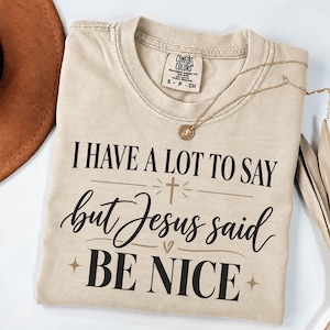 May include: A beige short-sleeve t-shirt with a Comfort Colors label at the neck. The front features black text that reads I HAVE A LOT TO SAY but Jesus said BE NICE, accented with a small cross, a heart, and star symbols in a tan color.