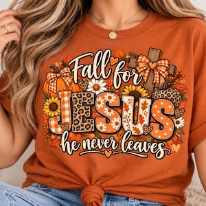 May include: A burnt orange t-shirt featuring a graphic print with the text Fall for JESUS he never leaves. The design includes a pumpkin, a wooden cross with a plaid bow, sunflowers, daisies, and autumn leaves in orange, brown, and white tones.