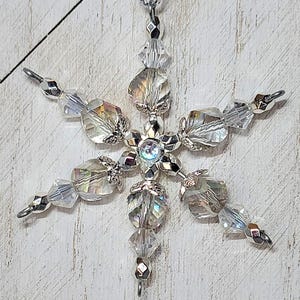 May include: A handcrafted snowflake ornament with clear, iridescent beads and silver-toned metal accents. The six-pointed star design features a central iridescent bead and is suspended by a silver cord. A decorative winter holiday item.