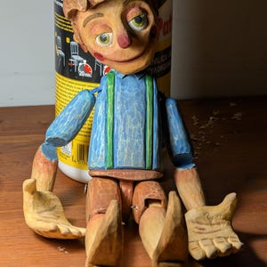 Può includere: Un burattino di legno con un viso scolpito, capelli castani e occhi verdi. La bambola indossa una camicia blu con bretelle verdi e arti in legno. Le mani della bambola sono aperte e le articolazioni sono visibili. Una bottiglia è sullo sfondo.