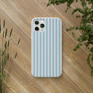 May include: A smartphone case with a vertical striped pattern in light blue and white. The case is on a wooden surface with green foliage in the background. The case has a camera module with three lenses.
