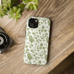 Puede incluir: Una funda de teléfono blanca con un estampado floral verde con rosas y hojas. La funda está sobre una superficie de madera, junto a una cámara y una planta.