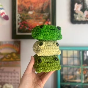 May include: Three handmade crochet frog plushies stacked on top of each other. The top and bottom frogs are a vibrant green, while the middle frog is a light green. Each frog has black button eyes and a smiling mouth.