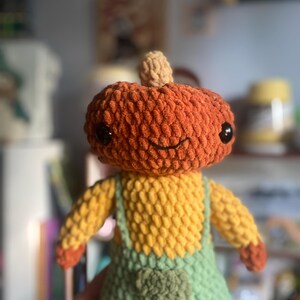 May include: A handmade crochet pumpkin doll with a smiling face, black eyes, and a tan stem. The doll wears yellow overalls with green bibs and a small pocket. The arms and hands are yellow and orange. The doll is a fall-themed decorative item.
