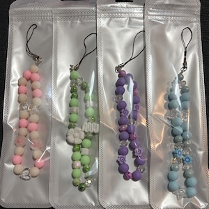 May include: Four beaded phone charms in clear plastic packaging. The charms feature various colours, including pink, green, purple, and blue. Each charm has a black lanyard and a mix of beads and decorative elements.