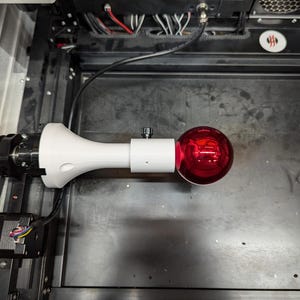 May include: A white and black camera with a red light attached to a white arm. The camera is mounted on a black surface.