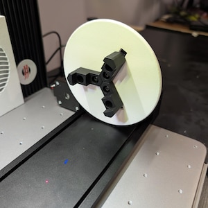 May include: A white circular platform with a black three-pronged rotating attachment. The platform is attached to a black metal base.