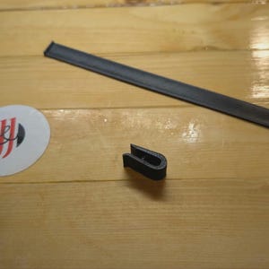 May include: A black leather strap and a U-shaped black object are displayed on a wooden surface. A white circular sticker with a red and black logo is also present. The items appear to be accessories or components.