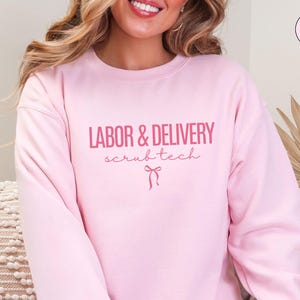 May include: Light pink sweatshirt with the words "LABOR & DELIVERY scrubtech" in pink script. A pink bow graphic is below the text. The sweatshirt is a solid light pink color.