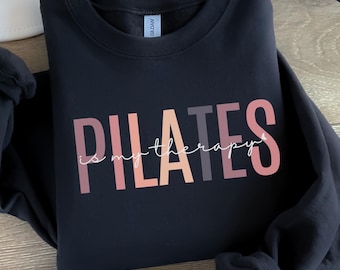 May include: A black crewneck sweatshirt featuring the word PILATES in large, multi-colored block letters in shades of pink, peach, and grey. The phrase is my therapy is written in a white cursive script across the center of the block letters.
