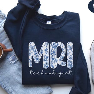 May include: Navy blue sweatshirt with the letters "MRI" in a white floral design. The word "technologist" is written in a script font below the letters. A coffee mug and a pair of gray sneakers are visible in the image.