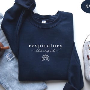 May include: Navy blue sweatshirt with the words "respiratory therapist" in white script, along with a white graphic of lungs. The sweatshirt is paired with light blue jeans, a brown mug, and gray sneakers.