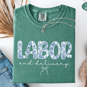 Op de afbeelding: Een lichtgroen t-shirt met de woorden "LABOR and delivery" in witte letters en blauwe bloemenprint. Een zilveren ketting met een hanger ligt op het shirt. Het shirt is op een wit oppervlak gevouwen naast een spijkerbroek.