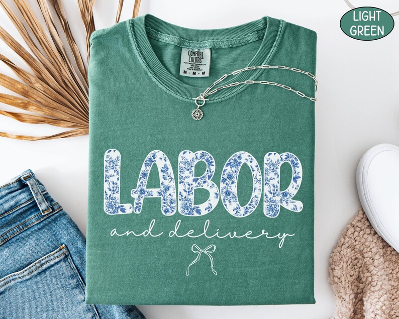 Op de afbeelding: Een lichtgroen t-shirt met de woorden "LABOR and delivery" in witte letters en blauwe bloemenprint. Een zilveren ketting met een hanger ligt op het shirt. Het shirt is op een wit oppervlak gevouwen naast een spijkerbroek.