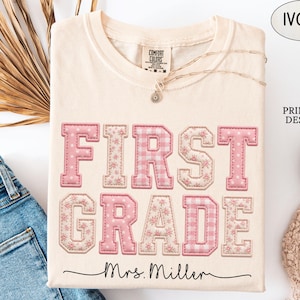 May include: An ivory short-sleeved t-shirt featuring the text FIRST GRADE in pink varsity-style letters with floral and gingham patterns. Below the text, the name Mrs. Miller is printed in black cursive script. The shirt has a crew neckline.